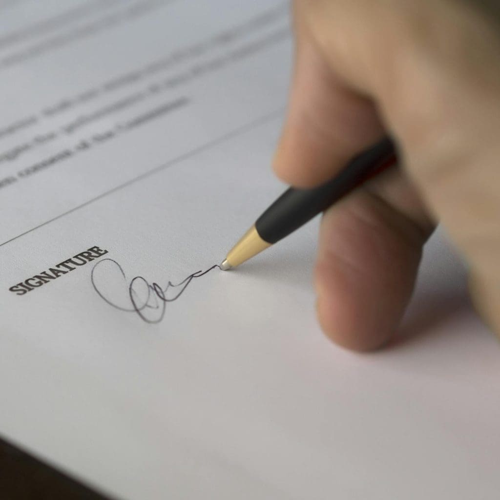 A hand signs a formal contract with a pen on a wooden desk.